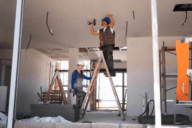 Worker on ladder using drill, another observing, amid scaffolding and materials.