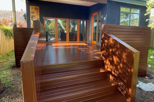 Newly built brown wooden deck with steps, railings, and large windows reflecting sunlight.