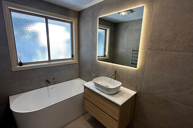 Modern bathroom with freestanding bathtub, wooden vanity, bowl sink, and illuminated mirror.