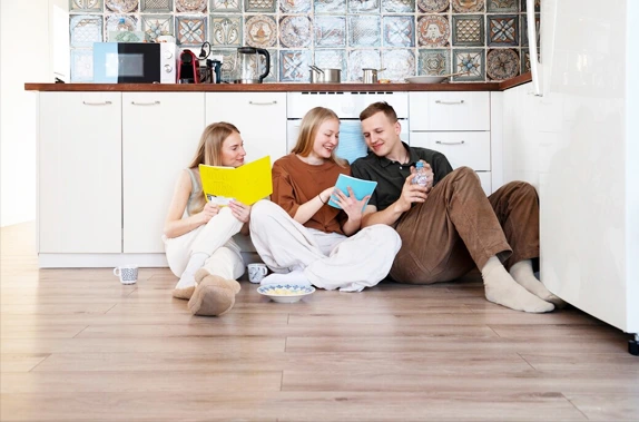 Three people sitting on the kitchen floor, reading colorful books, with mugs and a bowl of snacks.